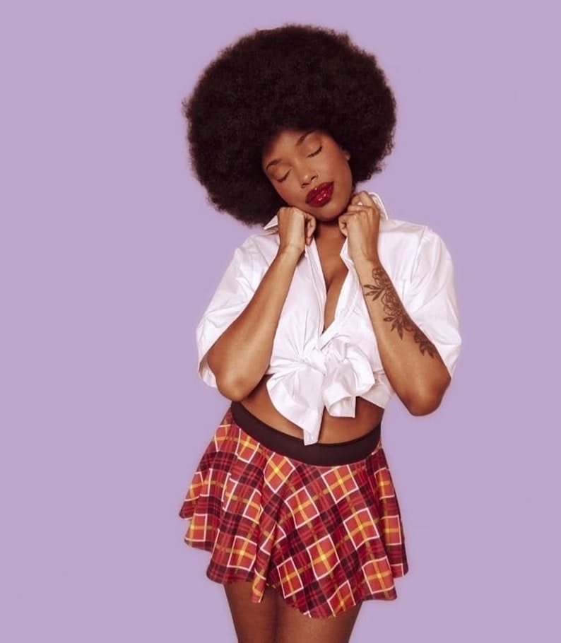 K&ouml;nnte beinhalten: Eine Frau mit Afrofrisur tr&auml;gt ein wei&szlig;es Hemd mit Kn&ouml;pfen, das in der Taille gebunden ist, und einen rot-schwarzen karierten Rock.