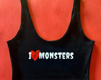 May include: Black crop top with the text "I ❤️ MONSTERS" in white. The heart is red with a stitched design. The top has a scoop neck and wide straps. The background is red.