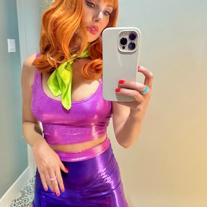 May include: A purple metallic crop top and matching mini skirt costume with a light purple trim. The outfit is paired with an orange wig, a green scarf, and a purple headband. The costume is reminiscent of a cartoon character.