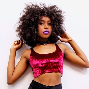 May include: A burgundy velvet crop top with a scoop neck. The top is paired with a black choker necklace with a small silver pendant. The model has dark curly hair and purple lipstick.
