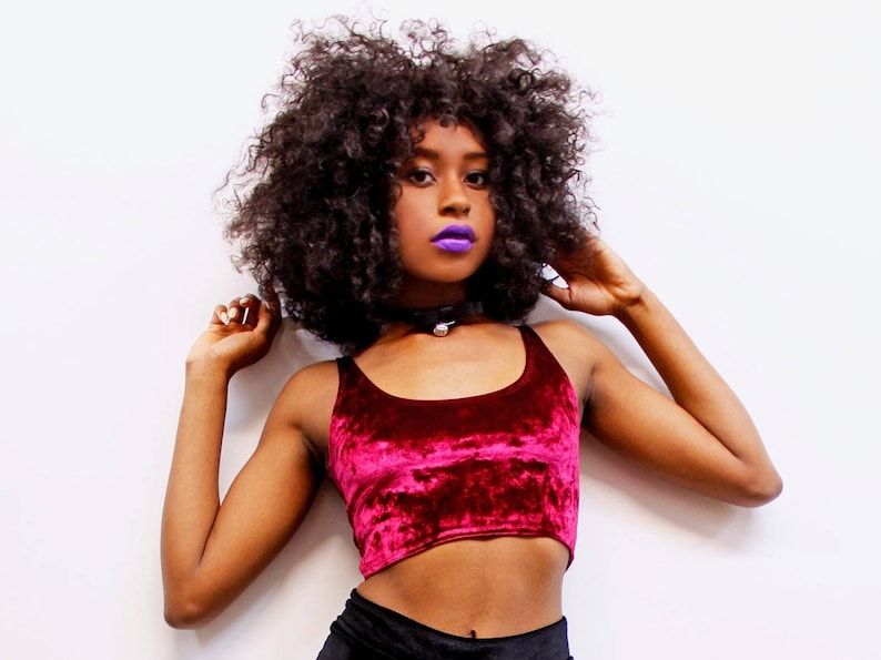 May include: A burgundy velvet crop top with a scoop neck. The top is paired with a black choker necklace with a small silver pendant. The model has dark curly hair and purple lipstick.