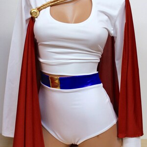 Puede incluir: Top blanco corto con un escote de ojo de cerradura y un detalle de cadena dorada. El top se combina con pantalones cortos blancos y una capa roja. Un cintur&oacute;n azul con una hebilla dorada completa el look.