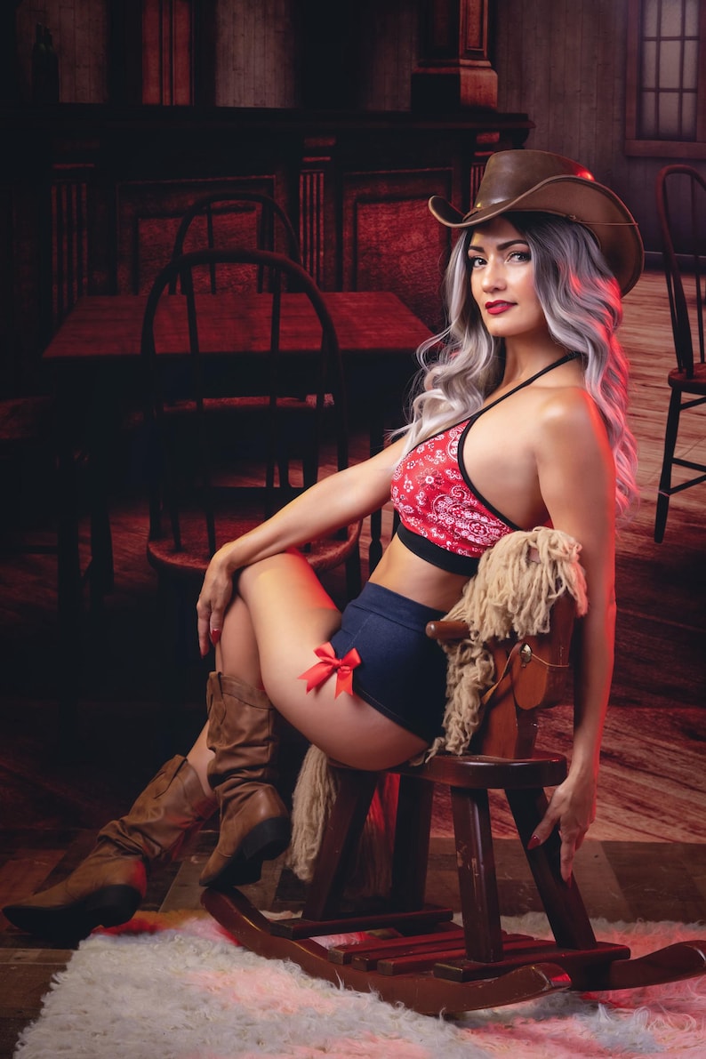 May include: A woman wearing a red and black bandana print halter top, blue denim shorts with a red bow, and brown cowboy boots. She is sitting on a rocking horse with a brown cowboy hat on her head. The background is a dark red and brown wooden bar.