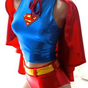 K&ouml;nnte beinhalten: Ein Superhelden-Kost&uuml;m mit einem blauen Oberteil mit Superman-Logo, roten Shorts, einem goldenen G&uuml;rtel und einem roten Umhang. Das Kost&uuml;m besteht aus gl&auml;nzendem Material und wird auf einer Schaufensterpuppe pr&auml;sentiert.