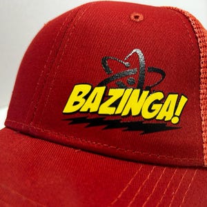 May include: Red and orange baseball cap with a black and yellow "Bazinga!" logo. The logo features a stylized atom symbol.