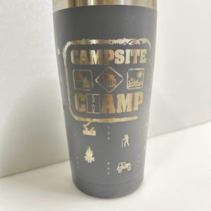 May include: Gray stainless steel tumbler with a gold inscription that reads "Campsite Champ" and features camping icons.