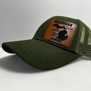 May include: A green and brown baseball cap with a mesh back. The front of the cap features a brown leather patch with the text "MICHIGAN" and an outline of the state of Michigan. The text on the patch reads "Pay the toll and go. Truck Township. Round About County. Zoning."