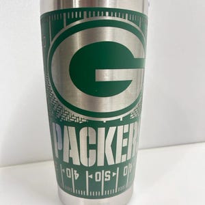 May include: A stainless steel tumbler with a green and silver Green Bay Packers logo. The tumbler has a ruler design around the bottom.