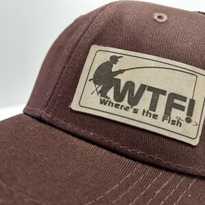 May include: A brown and tan baseball cap with a mesh back. The front of the cap features a brown patch with a silhouette of a person fishing and the text "WTF! Where's the Fish?"