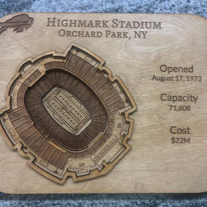 Può includere: Targa in legno con una rappresentazione dettagliata, tagliata al laser, dell'Highmark Stadium a Orchard Park, NY. Il layout dello stadio è ben definito. Il testo include nome, luogo, data di apertura, capacità di 71.608 e costo.