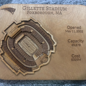 May include: A wooden plaque with a laser-cut representation of Gillette Stadium in Foxborough, MA. The stadium's design is finely carved, with text detailing the stadium's name, location, opening date, capacity (65,878), and cost.