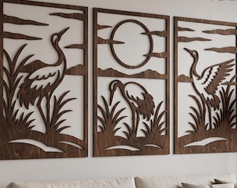 May include: A set of three rectangular dark brown wooden wall panels featuring laser-cut silhouettes of cranes standing in tall grass. The center panel includes a circular sun design, while the outer panels depict cranes in various poses.