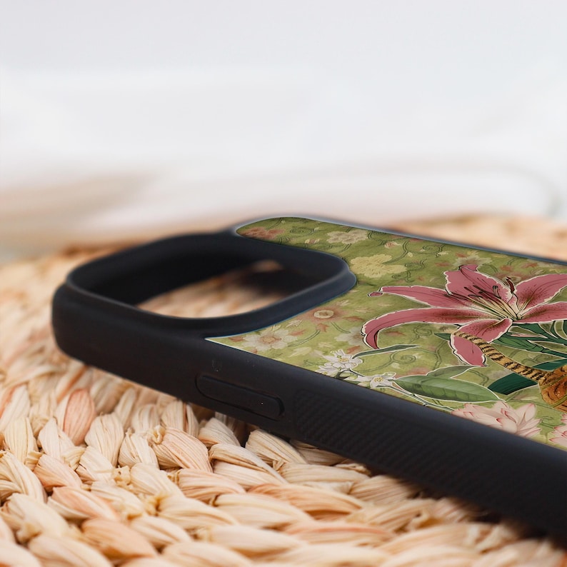 May include: Black phone case featuring a floral design with a pink lily and green leaves on a light green background. The case has a black border and is resting on a woven surface.