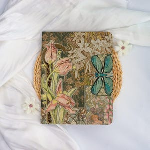 May include: A decorative journal with a floral design featuring pink tulips and a teal dragonfly. The cover has a dark green background with a collage of floral patterns and artwork. The journal is placed on a woven mat.
