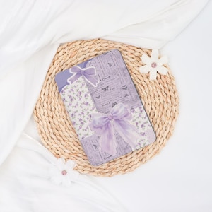 May include: A decorative rectangular box with a lavender floral and butterfly design, accented with lavender bows. The box is placed on a woven mat, with white fabric in the background.