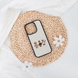 May include: A phone case with a black border and a white background covered in small black polka dots. The case features an illustration of three teddy bears wearing outfits. The case is resting on a woven mat.