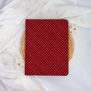 Peut inclure: Un cahier rouge avec un motif à pois blancs. Le cahier est posé sur un dessous de plat en osier et un tissu blanc. Deux fleurs blanches sont placées près du cahier.