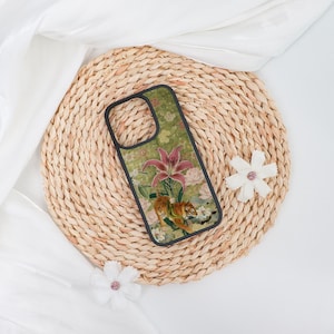 May include: A phone case featuring a floral and tiger design. The case has a black border and a green background with pink flowers and a tiger. The case is on a woven mat with white fabric and decorative flowers.