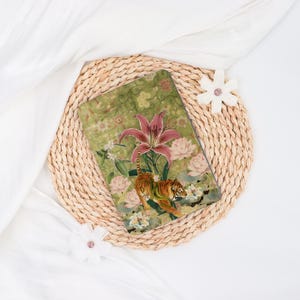 May include: A decorative tablet case featuring a tiger amidst pink lilies and other flowers on a green background. The case is set on a woven placemat with white fabric and flower accents.