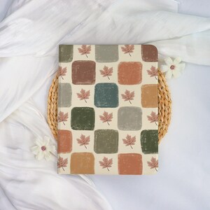 May include: A rectangular notebook with a cream-colored background featuring a grid pattern of squares in various muted colors, including brown, green, and orange. Each square has a small, pink maple leaf. The notebook is on a woven placemat.