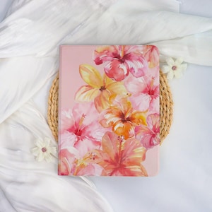 May include: A pink tablet case featuring a watercolor floral design of hibiscus flowers in shades of pink, orange, and yellow. The case is set on a woven placemat and white fabric.