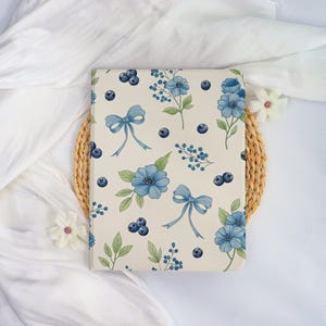 May include: A cream-colored notebook featuring a floral pattern with blue flowers, bows, blueberries, and green leaves. The notebook is placed on a woven mat and white fabric.