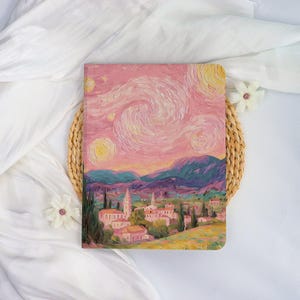 Könnte beinhalten: Ein Notizbuch mit einem Gemälde eines Dorfes unter einem wirbelnden Sternenhimmel in Rosa, Gelb und Blau. Das Kunstwerk ist im Stil von Van Gogh gehalten, mit sichtbaren Pinselstrichen und einer strukturierten Optik. Das rechteckige Notizbuch liegt auf einem geflochtenen Untersetzer.