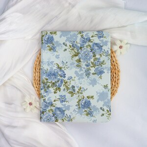 May include: A floral-patterned notebook featuring a light blue background with blue rose illustrations and green leaves. The notebook is resting on a woven circular placemat, with white fabric and decorative flowers in the background.
