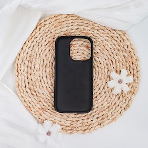 May include: A black phone case with a smooth finish rests on a round, woven straw mat. Two white fabric flowers with pink centers are placed near the case. The background includes white fabric.