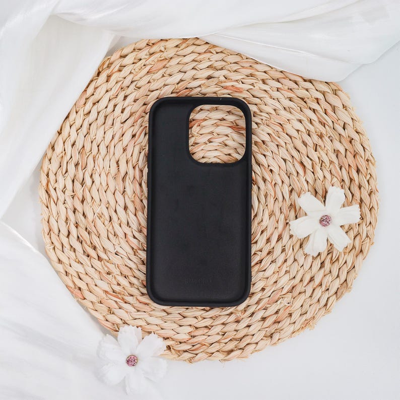 May include: A black phone case with a smooth finish rests on a round, woven straw mat. Two white fabric flowers with pink centers are placed near the case. The background includes white fabric.