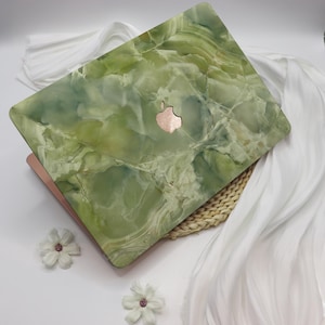 Könnte beinhalten: Grüne Marmor-Laptop-Haut mit einem silbernen Apple-Logo. Die Haut liegt auf einer weißen Oberfläche mit einer geflochtenen Strohmatte darunter.