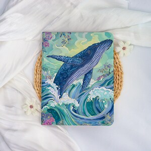May include: A journal with a vibrant illustration of a blue whale breaching the ocean surface. The cover features a detailed design with swirling waves, floral accents, and a gradient sky. The color palette includes shades of blue, green, and purple.