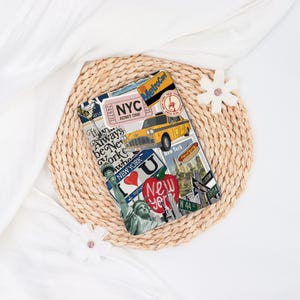 May include: A rectangular New York City-themed notebook featuring a yellow taxi, the Statue of Liberty, and the text "NYC". The notebook is covered in various stickers and images associated with New York City.