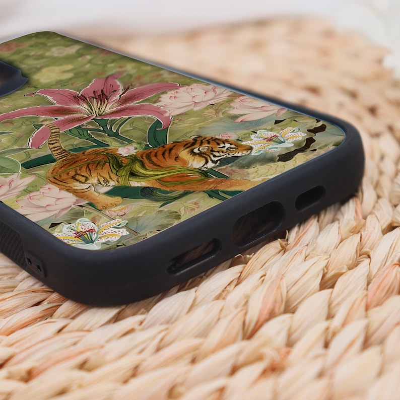 May include: Black phone case featuring a detailed illustration of a tiger amidst pink lilies and green foliage. The design has a vintage, artistic style. The case is made of a durable material and has a sleek, modern design.