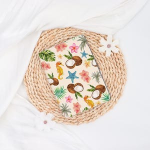 May include: A rectangular notebook with a tropical design featuring coconuts, seahorses, starfish, and flowers. The notebook is set on a woven placemat with white fabric and decorative flowers.