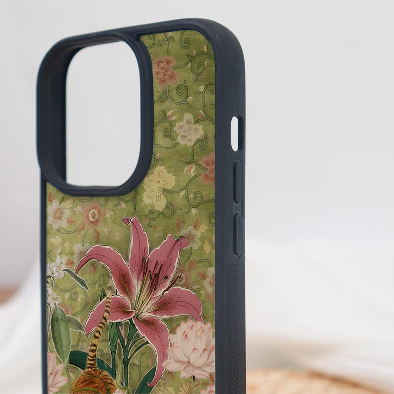 May include: A phone case with a floral design featuring a pink lily and other flowers on a green background. The case has a dark blue border and a cutout for the camera and buttons. The design includes a tiger.