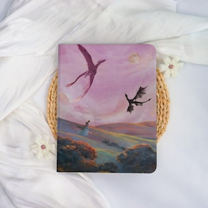 May include: A fantasy-themed journal featuring a painting of dragons soaring across a pink and purple sky. A figure in a flowing gown walks on a hillside. A full moon is visible in the background. The journal is rectangular.