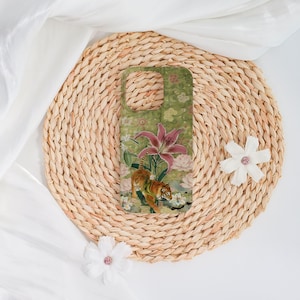 May include: A green phone case featuring a tiger illustration with pink lilies and white flowers. The case is set on a woven straw mat with white fabric and decorative flowers.