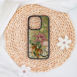May include: A phone case with a black border featuring a floral and animal design. The case has a green background with a tiger, pink lilies, and white flowers. The case is set on a woven, circular placemat.