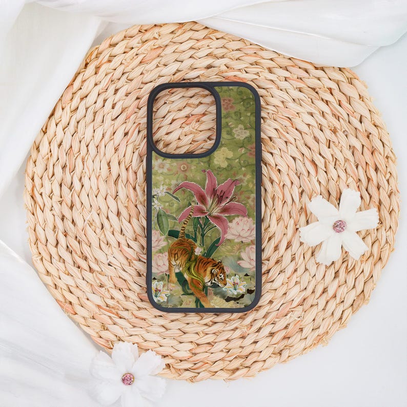 May include: A phone case with a black border featuring a floral and animal design. The case has a green background with a tiger, pink lilies, and white flowers. The case is set on a woven, circular placemat.