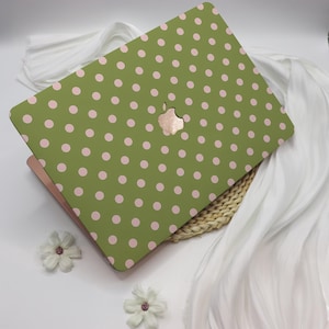 May include: A green laptop case with a pink polka dot pattern. The case has a cutout for the Apple logo. The laptop case is on a white surface with white fabric and two white flowers.