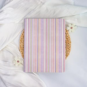 May include: A rectangular notebook with a pastel striped pattern in shades of pink, purple, and yellow. The notebook is resting on a woven placemat, with white fabric and decorative flowers in the background.