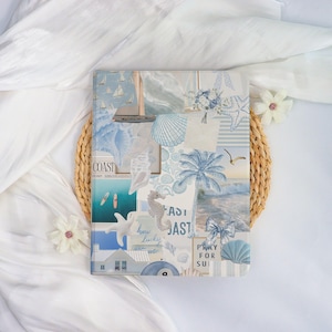 May include: A notebook with a collage design featuring nautical elements in shades of blue and white. The design includes sailboats, seashells, palm trees, and the text "COAST". The notebook is placed on a woven placemat and white fabric.