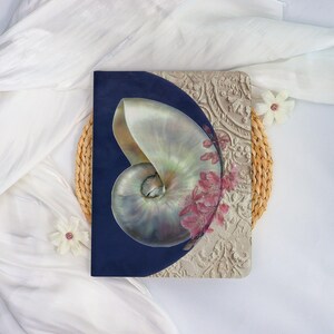 May include: A decorative notebook featuring a nautilus shell design. The shell is painted with iridescent colors and set against a navy blue background. Pink floral accents and a textured, beige patterned section complete the design.