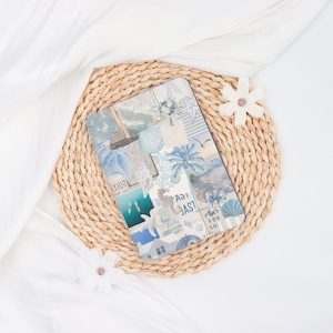 May include: A rectangular tablet case with a collage design featuring coastal elements in shades of blue and white. The design includes images of sailboats, seashells, and palm trees. The case rests on a woven placemat, with white fabric and decorative flowers.