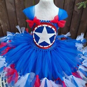 May include: A blue, red, and white tutu dress with a Captain America shield design on the bodice. The dress features a star-shaped emblem and red bows on the shoulders. The skirt is layered with blue tulle and accented with red and white tulle.