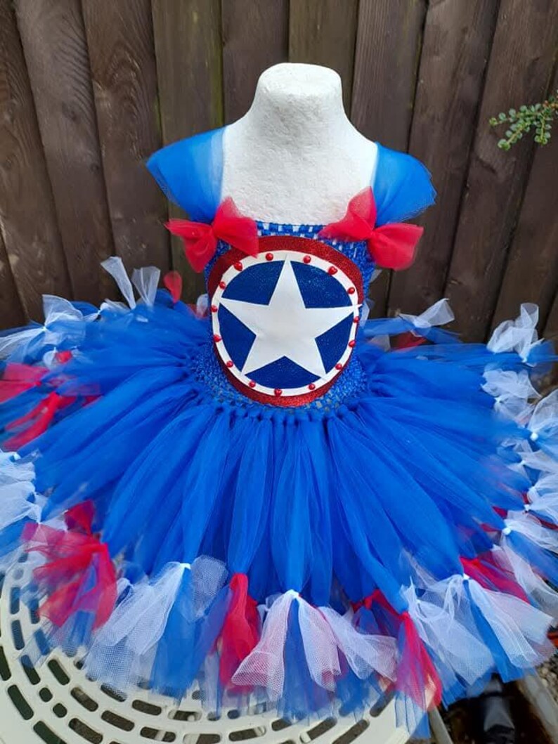 May include: A blue, red, and white tutu dress with a Captain America shield design on the bodice. The dress features a star-shaped emblem and red bows on the shoulders. The skirt is layered with blue tulle and accented with red and white tulle.