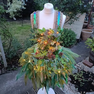 Puede incluir: Un vestido de hada verde y marrón en un maniquí blanco. El vestido está adornado con hojas artificiales, piñas y luces de hadas. Las correas están adornadas con lentejuelas iridiscentes. La falda es de tela verde en capas.