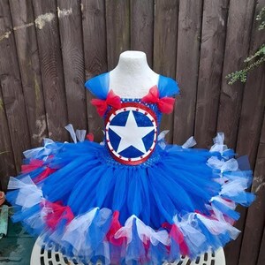 May include: A blue, red, and white tutu dress with a Captain America shield design. The dress features a blue bodice with a shield emblem and a layered tulle skirt. Red bows adorn the shoulders. The dress is displayed on a mannequin.