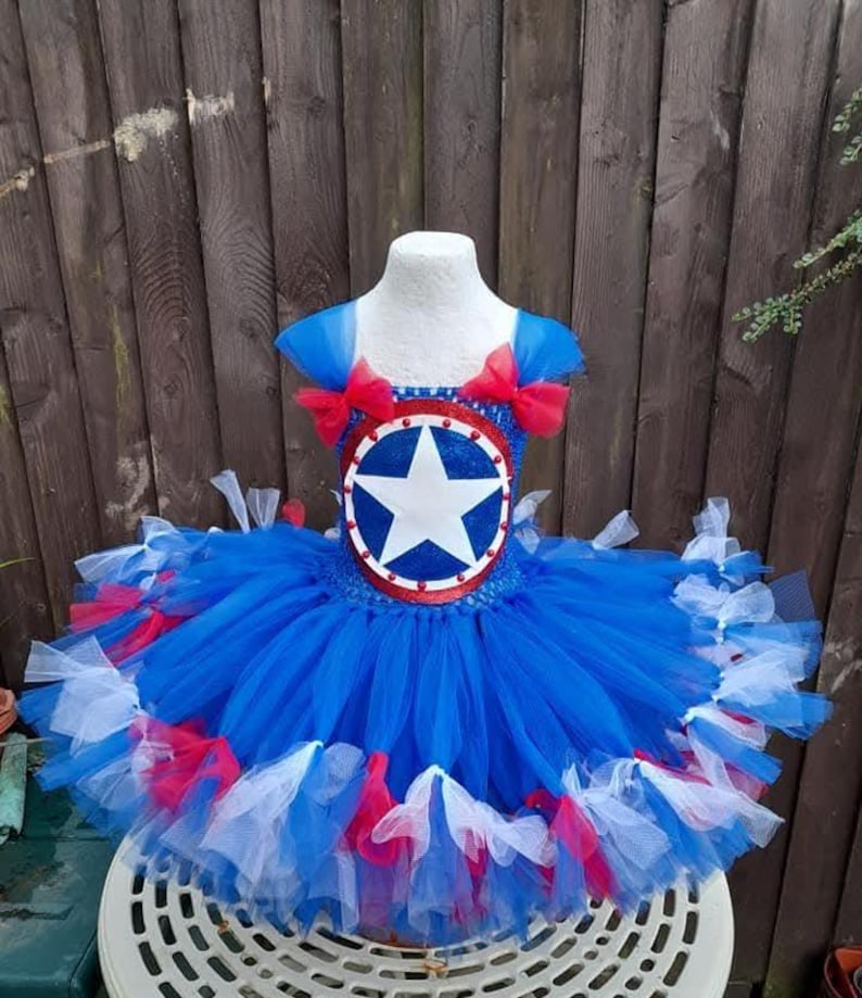 May include: A blue, red, and white tutu dress with a Captain America shield design. The dress features a blue bodice with a shield emblem and a layered tulle skirt. Red bows adorn the shoulders. The dress is displayed on a mannequin.